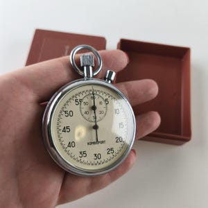 May include: A silver-toned analog stopwatch with a cream-colored face, black numerals, and a sub-dial. The dial features the text KOMBISPORT. It includes a top winding crown and a side button, accompanied by a separate brown plastic storage case.