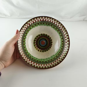 May include: A round ceramic bowl with a white interior and a decorative rim. The rim features a brown zig-zag pattern, green dots, and a central design with concentric circles in brown, blue, and green. The bowl is held by a hand.