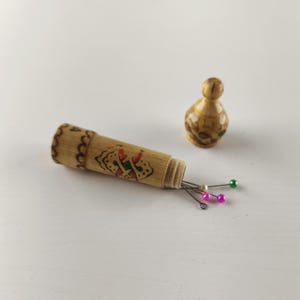 Puede incluir: Un peque&ntilde;o portagujas cil&iacute;ndrico de bamb&uacute; con un dise&ntilde;o decorativo y una tapa a juego separada. El portagujas contiene varias agujas con cabezas coloridas. Los art&iacute;culos est&aacute;n sobre una superficie blanca.