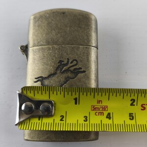 May include: Antique brass-colored lighter with a rodeo rider and bull design. The lighter is approximately 2 inches long. The tape measure shows the lighter's length in inches and centimeters.