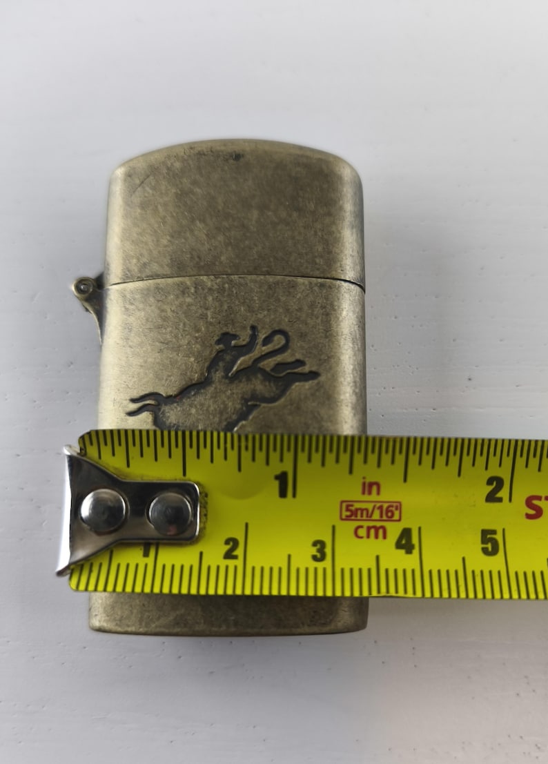 May include: Antique brass-colored lighter with a rodeo rider and bull design. The lighter is approximately 2 inches long. The tape measure shows the lighter's length in inches and centimeters.