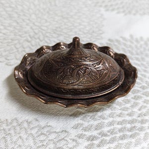 Może przedstawiać: Mały, antyczny talerz w kolorze miedzi z pokrywką. Talerz ma falisty brzeg i skomplikowane wzory kwiatowe wytrawione zarówno na pokrywce, jak i na podstawie. Pokrywka ma mały uchwyt na górze dla łatwej obsługi. Talerz jest prawdopodobnie używany do serwowania masła lub innych drobnych przedmiotów.