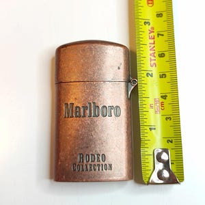 May include: A copper-toned Marlboro Rodeo Collection lighter. The rectangular lighter has rounded edges and the brand name and collection name are displayed. A yellow tape measure indicates the lighter is approximately 7.6 cm in height.