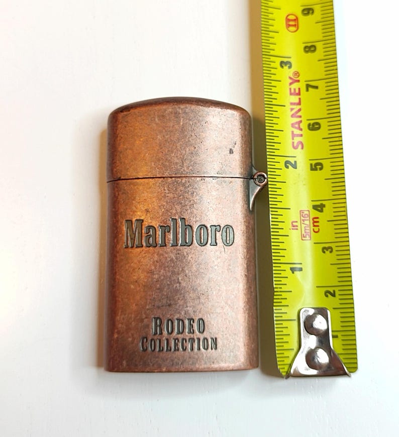 May include: A copper-toned Marlboro Rodeo Collection lighter. The rectangular lighter has rounded edges and the brand name and collection name are displayed. A yellow tape measure indicates the lighter is approximately 7.6 cm in height.