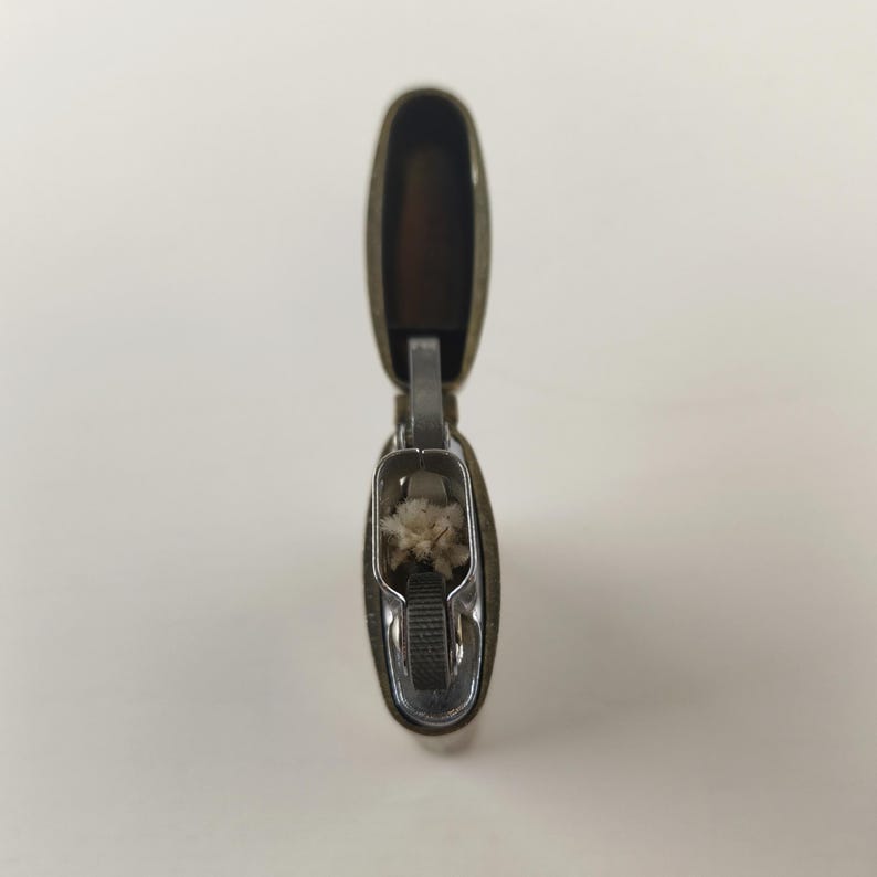 May include: An open, antique-style lighter with a brass-colored exterior and a silver-colored interior. The lighter's lid is raised, revealing the wick and interior components. The lighter is on a white background.