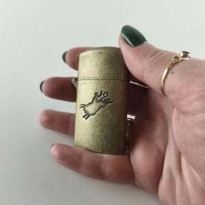 May include: A vintage-style brass lighter with a detailed embossed image of a pig. The lighter has a rectangular shape with rounded edges and a hinged lid. The pig design is in a darker shade, contrasting with the lighter brass color.