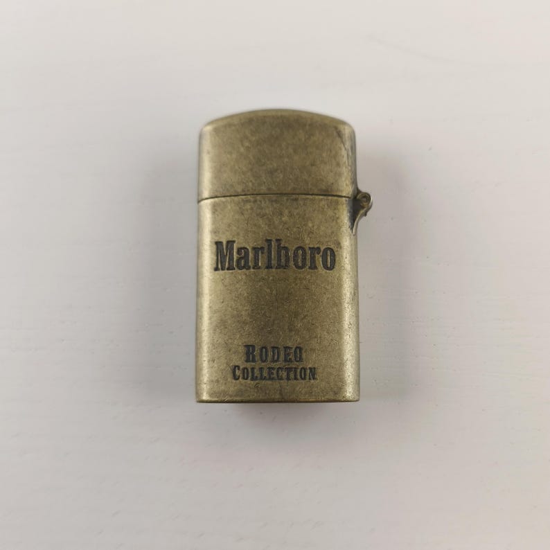 May include: A vintage brass-colored lighter with the word "Marlboro" and "Rodeo Collection" engraved on the front. The lighter has a rectangular shape with rounded edges and a hinged lid. The lighter is a collectible item.