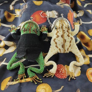 以下が含まれることがあります： 2つのビーズで作られたカエル型の小銭入れ。1つは緑と黒、もう1つはクリームとゴールドです。どちらも銀色の金属製の留め金が付いており、キノコの模様が入った濃い青色の生地の上にあります。カエルは詳細なビーズ細工と伸びた手足を持っています。