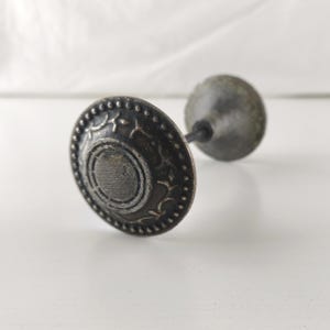 May include: A round metal cabinet knob with a dark, weathered finish. The front face features a raised circular center surrounded by a decorative border of small dots and etched leaf-like patterns. A thin metal post extends from the back.