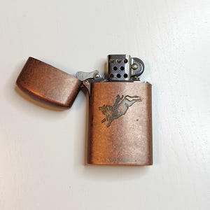 May include: Copper-coloured metal lighter with an open lid. The lighter has a detailed embossed design of a person riding a bull. The lighter is rectangular with rounded edges and a hinged lid.