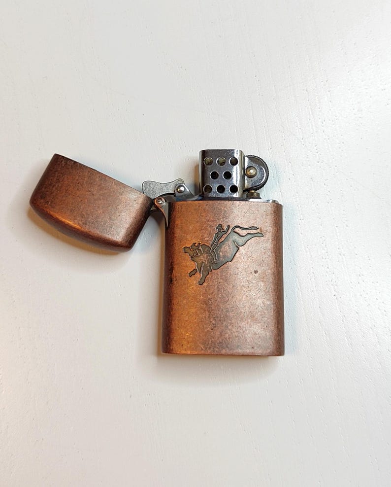 May include: Copper-coloured metal lighter with an open lid. The lighter has a detailed embossed design of a person riding a bull. The lighter is rectangular with rounded edges and a hinged lid.