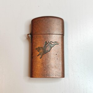 May include: A copper-toned lighter with a hinged lid. The front of the lighter features a detailed illustration of a person riding a bull. The lighter has a metallic finish and a small lever on the side.