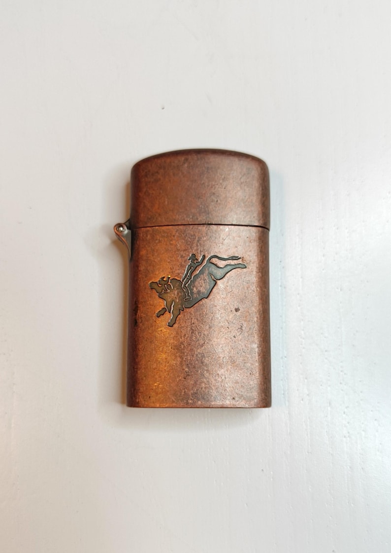 May include: A copper-toned lighter with a hinged lid. The front of the lighter features a detailed illustration of a person riding a bull. The lighter has a metallic finish and a small lever on the side.