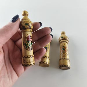 Puede incluir: Tres objetos decorativos de madera con acabado marr&oacute;n claro. Cada objeto tiene una parte superior redondeada y un cuerpo cil&iacute;ndrico con dise&ntilde;os florales pintados en rojo, verde y amarillo. La palabra "BUL" es visible en un objeto.