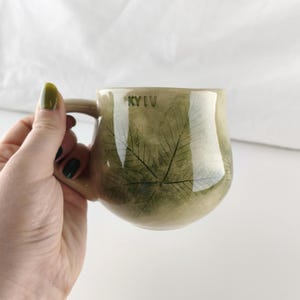 May include: A round ceramic mug with a glossy finish, featuring a muted green and beige mottled glaze. The surface displays a dark green leaf imprint pattern and the word KYIV stamped in capital letters near the top rim, with a curved handle.