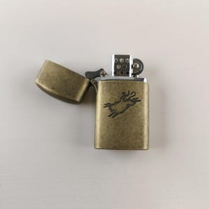 May include: An antique brass-colored lighter with an open lid. The lighter features a detailed engraving of a horse on its front. The lighter is rectangular and has a hinged lid. The lighter is on a white surface.