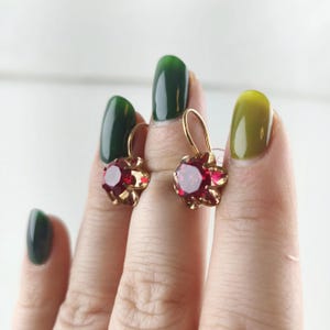 Puede incluir: Un par de pendientes de oro con piedras redondas de color rojo oscuro facetadas en un marco de metal inspirado en una flor. Cada pendiente tiene un cierre de gancho curvo y una gema central de color carmesí vibrante.