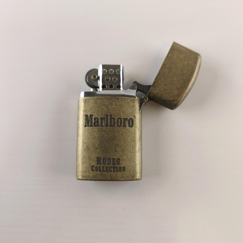 May include: A vintage-style, brass-colored lighter with the word "Marlboro" and "Rodeo Collection" printed on the front. The lid is open, revealing the interior. The lighter is on a white surface.