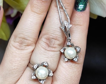 May include: A silver jewellery set comprising a ring and a pendant necklace. Each piece features a flower design with a central pearl and small, clear gemstones. The necklace has a fine chain.
