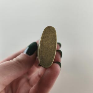 May include: A close-up of a vintage-style, oval-shaped lighter with a brass finish. The lighter has a textured surface and a slightly worn appearance, suggesting age and use. The lighter is held in a hand with dark green nail polish.