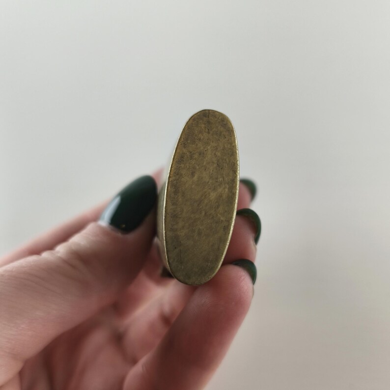 May include: A close-up of a vintage-style, oval-shaped lighter with a brass finish. The lighter has a textured surface and a slightly worn appearance, suggesting age and use. The lighter is held in a hand with dark green nail polish.