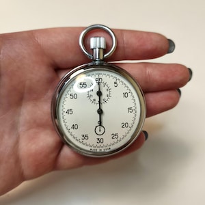 May include: A silver-toned mechanical stopwatch with a round metal case and a circular ring at the top. The white dial features black numerals from 5 to 60, a smaller sub-dial, and black hands. The text MADE IN USSR is printed at the bottom.