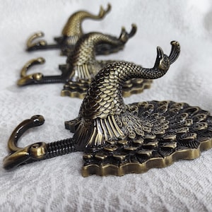 Puede incluir: Tres ganchos de metal de color dorado antiguo con forma de pavo real y un diseño detallado. Los ganchos tienen un aspecto vintage.