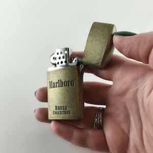 May include: A vintage brass-colored lighter with the word "Marlboro" and "Rodeo Collection" printed on the front. The lighter's top is open, revealing the internal mechanism. The lighter is held in a hand.