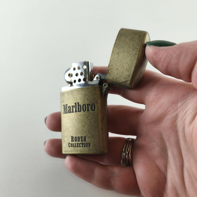 May include: A vintage brass-colored lighter with the word "Marlboro" and "Rodeo Collection" printed on the front. The lighter's top is open, revealing the internal mechanism. The lighter is held in a hand.