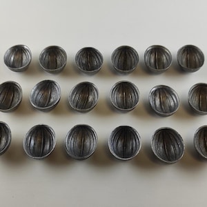 May include: Arrangement of eighteen small, metallic-looking, half-sphere objects with vertical grooves. The objects are arranged in three rows of six on a white surface. The objects have a dark, aged appearance.