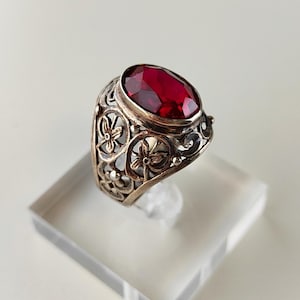Pode incluir: Anel de prata com uma grande pedra preciosa vermelha escura de corte oval. O anel apresenta um design ornamentado e vazado com detalhes florais e de rolagem. A pedra preciosa é definida em uma configuração de moldura. O anel é exibido em um suporte retangular transparente.