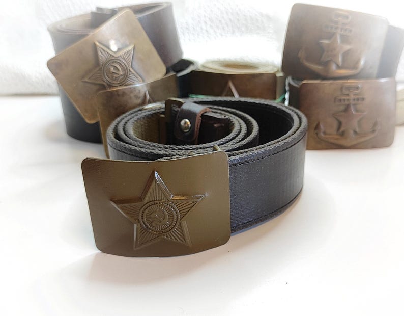 K&ouml;nnte beinhalten: Drei Milit&auml;rg&uuml;rtel mit Metallschnallen, die ein Stern- und Ankerdesign aufweisen. Die G&uuml;rtel sind braun und gr&uuml;n mit einem gewebten Stoffband.