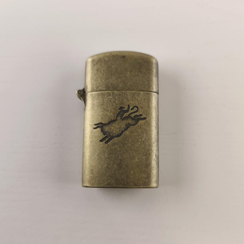 May include: A vintage-style brass lighter with a rectangular shape. The lighter features a detailed engraving of a leaping animal on the front. The brass has a slightly aged appearance, giving it a classic, antique feel.