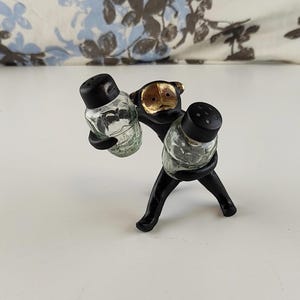 Puede incluir: Un juego de salero y pimentero negro con un diseño caprichoso de mono. El mono sostiene dos saleros de vidrio transparente con tapas negras. La cara del mono tiene detalles dorados. El juego está sobre una superficie blanca.