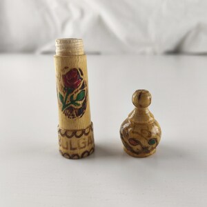 Puede incluir: Un peque&ntilde;o recipiente de madera con tapa extra&iacute;ble. El recipiente es cil&iacute;ndrico y presenta una rosa roja pintada con hojas verdes. La base del recipiente tiene la palabra "BULGA" tallada. La tapa tiene forma peque&ntilde;a y redondeada.