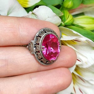 Könnte beinhalten: Ein silberner Ring mit einem ovalen, facettierten, leuchtend rosa Edelstein. Der Ring hat ein verziertes Filigran-Design um den Edelstein und an den Seiten. Im Hintergrund befinden sich weiße und gelbe Blütenblätter und grüne Blätter.