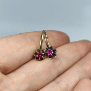 Può includere: Un paio di orecchini a fiore dorati con centri di pietre preziose rosse. Un orecchino ha un contorno floreale nero, l'altro dorato. Gli orecchini sono piccoli e delicati, con un semplice design a gancio.