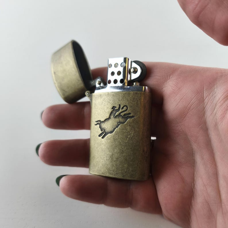 May include: A vintage-style brass lighter with an open lid. The lighter features a detailed embossed image of a leaping animal. The lighter is held in a hand, showcasing its size and design.