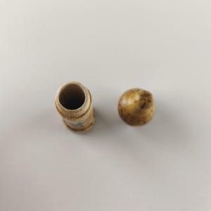 Puede incluir: Un peque&ntilde;o recipiente cil&iacute;ndrico de madera marr&oacute;n claro con una tapa redondeada. El recipiente tiene un interior hueco y una superficie ligeramente texturizada. La tapa es de un color similar y tiene una forma lisa y redondeada. Los art&iacute;culos est&aacute;n sobre una superficie blanca.