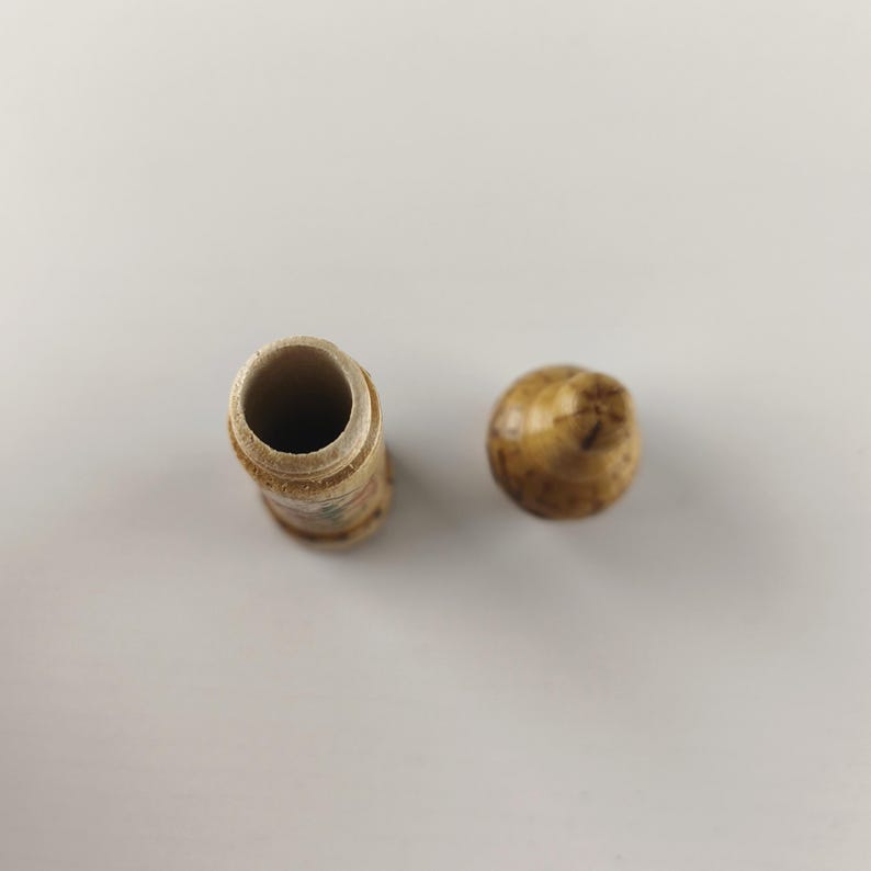 Puede incluir: Un peque&ntilde;o recipiente cil&iacute;ndrico de madera marr&oacute;n claro con una tapa redondeada. El recipiente tiene un interior hueco y una superficie ligeramente texturizada. La tapa es de un color similar y tiene una forma lisa y redondeada. Los art&iacute;culos est&aacute;n sobre una superficie blanca.