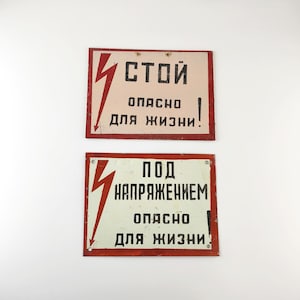 Op de afbeelding: Twee rechthoekige metalen borden met rode randen en bliksemgraphics. Het bovenste bord heeft de tekst "СТОЙ ОПАСНО ДЛЯ ЖИЗНИ!" en het onderste bord "ПОД НАПРЯЖЕНИЕМ ОПАСНО ДЛЯ ЖИЗНИ".