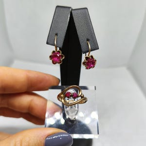 Op de afbeelding: Een goudkleurige sieradenset met een ring en oorbellen. De oorbellen en ring hebben elk een bloemmotief met helderrode edelstenen. De oorbellen zijn van het druppeltype en de ring heeft een uniek wervelontwerp.