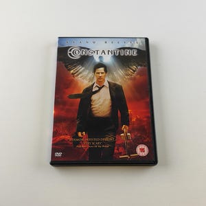 Op de afbeelding: DVD-hoes van de film "Constantine" met Keanu Reeves. De cover toont Reeves in een pak, met een wapen, tegen een vurige achtergrond, met de titel van de film prominent.