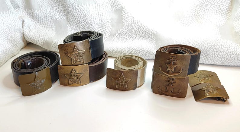 K&ouml;nnte beinhalten: F&uuml;nf Lederg&uuml;rtel mit verschiedenen Metallschnallen. Die Schnallen zeigen einen Stern, einen Hammer und eine Sichel sowie einen Anker. Die G&uuml;rtel sind braun und schwarz.