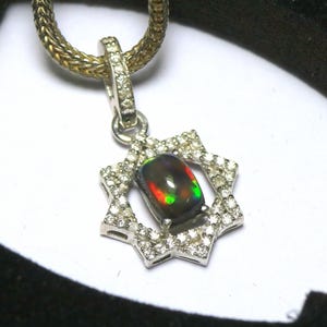 May include: A silver pendant featuring a black opal gemstone, set within a star-shaped frame of small, clear gemstones. The opal exhibits flashes of red, green, and yellow. The pendant is suspended from a silver chain.