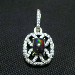 May include: A silver-toned pendant featuring a rectangular black opal with vibrant red, green, and blue flashes. The centre stone is surrounded by an intricate, openwork frame encrusted with small, clear, sparkling stones and a matching bail.