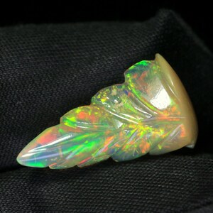 May include: A carved opal gemstone shaped like a leaf, featuring a translucent base with vibrant flashes of green, orange, and yellow play-of-color throughout the surface. The gemstone has a smooth, polished finish and a pointed tip.