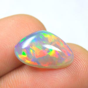May include: A smooth, teardrop-shaped opal gemstone featuring a translucent base with vibrant flashes of red, orange, yellow, and green color play across its polished surface. The stone exhibits a high-domed cabochon cut with a reflective finish.
