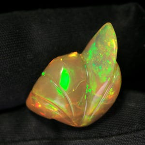 May include: A carved, translucent opal gemstone featuring a warm, peach-toned base color with vibrant, iridescent flashes of neon green and orange light. The stone is shaped with smooth, curved ridges that resemble a stylized leaf or petal form.