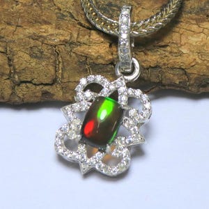 May include: A silver pendant featuring a rectangular black opal with vibrant green and red flashes, set within a decorative frame encrusted with small clear stones. The pendant hangs from a silver chain, with a stone-set bail connecting the two pieces.